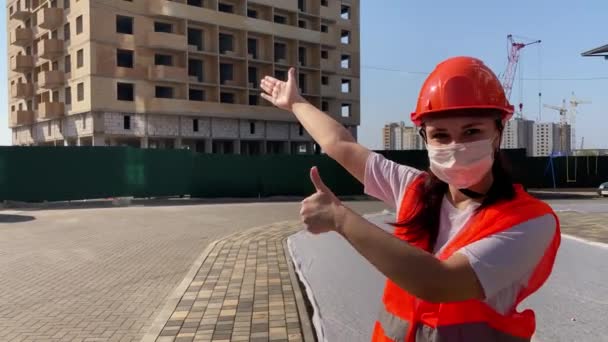 Female construction worker in overalls and in medical mask pointing at house under construction. — Stock Video