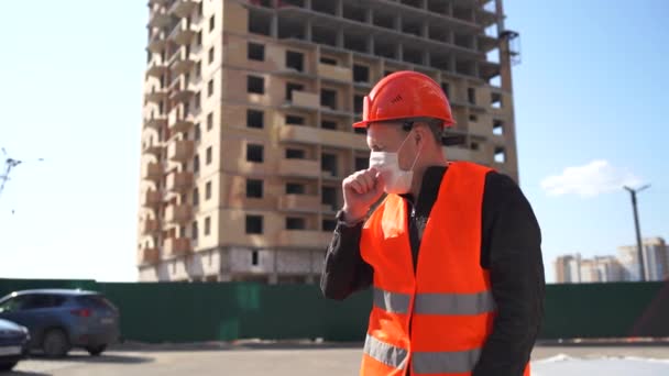 Мужчина строитель в комбинезоне и медицинская маска кашляет на фоне строящегося дома. Концепция угрозы заражения . — стоковое видео