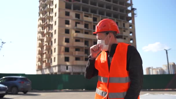 Muž stavební dělník v montérkách a lékařské masce kašlání na pozadí domu ve výstavbě. Pojetí hrozby infekce. — Stock video