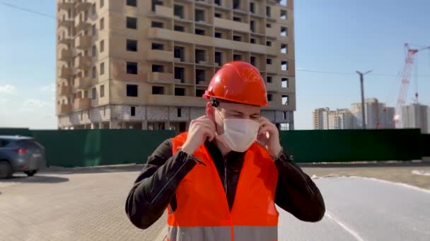 Мужчина строитель в комбинезоне снимает медицинскую маску с лица на строительной площадке — стоковое видео