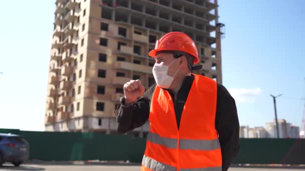 Мужчина строитель в комбинезоне и медицинская маска кашляет на фоне строящегося дома. Концепция угрозы заражения . — стоковое видео
