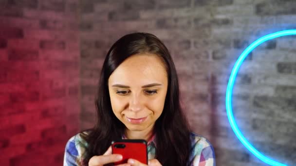 Jovem mulher bonita fica com telefone celular em mãos na parede de tijolo de fundo com lâmpadas brilhantes. Retrato de mulher encantadora lançando através de várias notícias no telefone celular contra a parede iluminada . — Vídeo de Stock