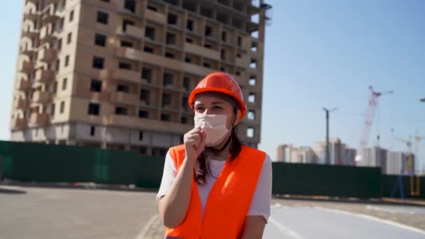 Eine Bauarbeiterin in Overalls und medizinischer Maske hustet vor dem Hintergrund eines im Bau befindlichen Hauses in die Faust. Konzept der Bedrohung durch Coronavirus-Epidemie. — Stockvideo