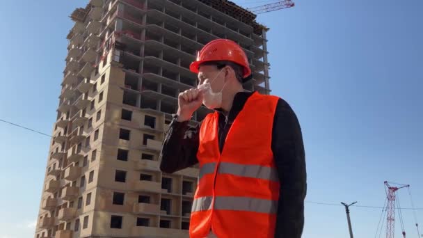 Männlicher Bauarbeiter in Overalls und medizinischer Maske hustet vor dem Hintergrund eines im Bau befindlichen Hauses. Konzept der Infektionsgefahr. — Stockvideo