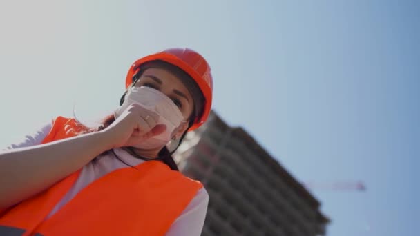 Kvinnlig byggarbetare i overaller och medicinsk mask hosta i knytnäve på bakgrund av hus under konstruktion. Begreppet hot om infektion med coronavirus. — Stockvideo