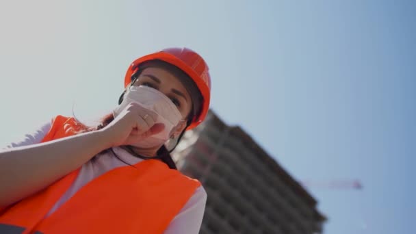 Ženská stavební dělnice v montérkách a s maskou kašlající v pěst na pozadí domu ve výstavbě. Koncepce hrozby nákazy koronavirem. — Stock video