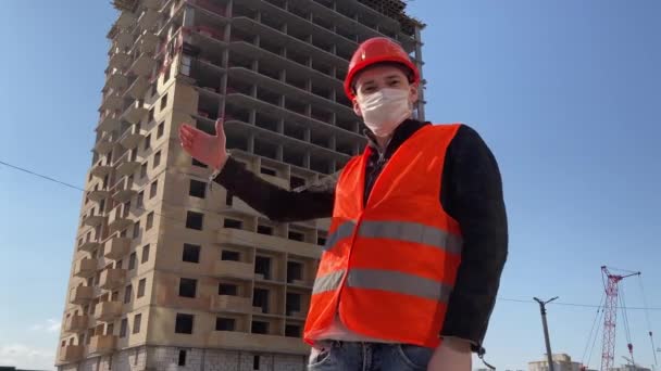 Homem trabalhador da construção civil em macacão e em máscara médica apontando para casa em construção . — Vídeo de Stock