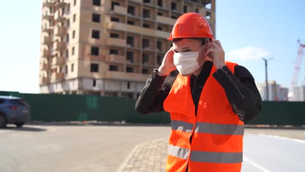 Mann Bauarbeiter in Overalls setzt medizinische Maske auf Gesicht vor Hintergrund von Haus im Bau. — Stockvideo