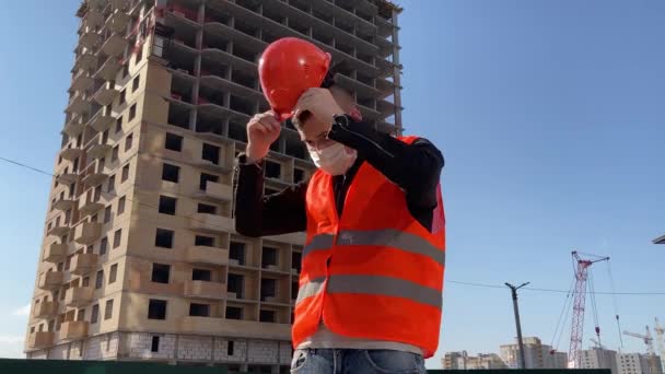 İnşaat halindeki evin arka planında tıbbi maskeli ve tulumlu erkek inşaat işçisinin portresi.. — Stok video