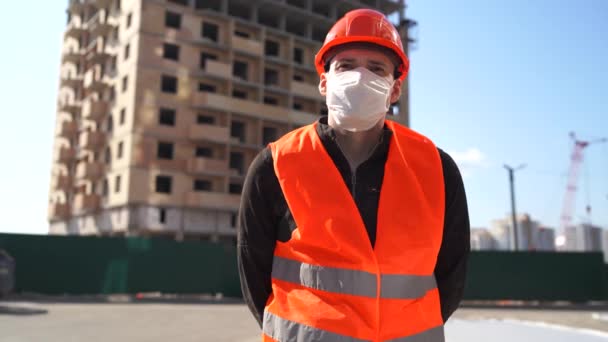 Portrét muže stavebního dělníka v lékařské masce a kombinézách na pozadí domu ve výstavbě. — Stock video