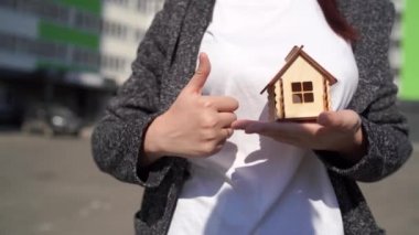 Elinde küçük ahşap bir ev tutan bir kadın ve diğer elinde inşa edilmiş bir evin arka planında baş parmağını gösteren bir kadın. Yeni daire satın alma kavramı.