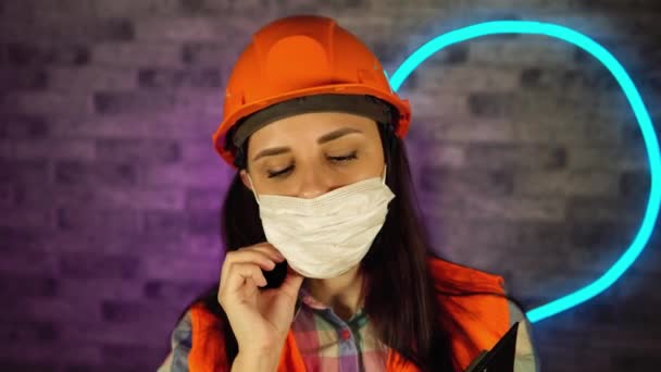 Vrouw in overall corrigeert medisch masker op haar gezicht, staat met klembord, bereidt zich voor op online werk tegen verlichte muur. Concept van dreiging van een besmetting met het coronavirus. — Stockvideo