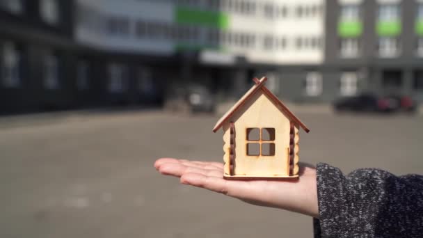Zblízka malý dřevěný dům v ženské ruce na pozadí postaveného domu. Koncept nákupu nového bytu. — Stock video