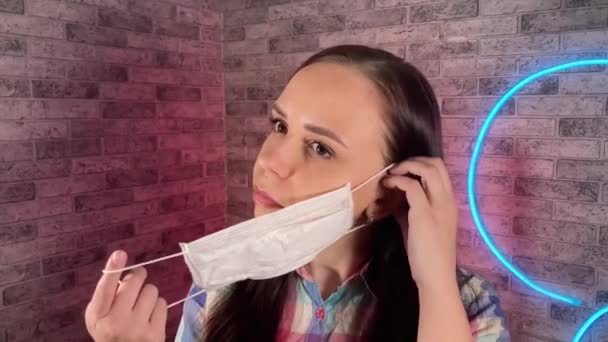 Πορτρέτο της ελκυστικής γυναίκας βάζοντας ιατρική μάσκα στο πρόσωπο κατά φωτισμένο τοίχο στο σπίτι. Κοντινό πλάνο των γυναικών που προστατεύουν τον εαυτό σας από ασθένειες. Έννοια της απειλής επιδημικής λοίμωξης από κορωνοϊό. — Αρχείο Βίντεο