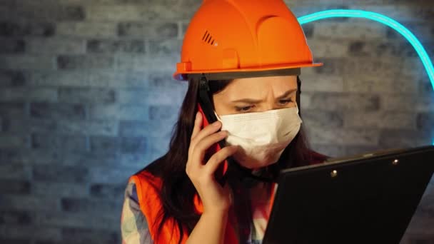 Mujer en overol y máscara médica hablando por teléfono inteligente y mirando en portapapeles contra la pared iluminada. Trabajadora de la construcción resolviendo preguntas sobre el proyecto empresarial, aislada en casa . — Vídeo de stock