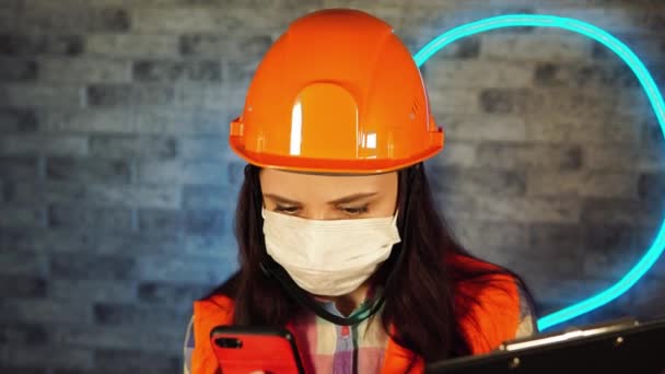 Mujer en mono y máscara médica mirando en el teléfono inteligente y portapapeles contra la pared iluminada. Trabajadora de la construcción revisando proyecto de negocio, siendo aislada en casa . — Vídeo de stock