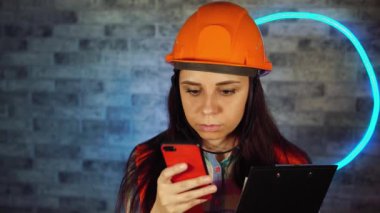 Tulumlu ve tıbbi maskeli bir kadın akıllı telefondan ışıklı duvara karşı pano ile sohbet ediyor. İş projesiyle ilgili soruları çözen, evde izole edilmiş kadın inşaat işçisi..