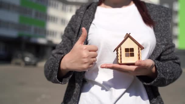 Close-up van de vrouw die kleine houten huis in de hand en andere hand tonen duim omhoog op de achtergrond van gebouwde huis. Concept van de aankoop van een nieuw appartement. — Stockvideo