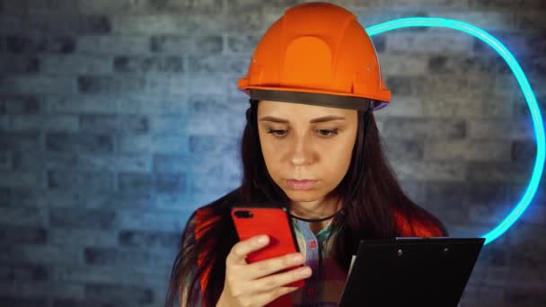 Woman in overalls and medical mask flipping chat in smartphone with clipboard against illuminated wall. Female construction worker solving questions about business project, being isolated at home. — Stock Video