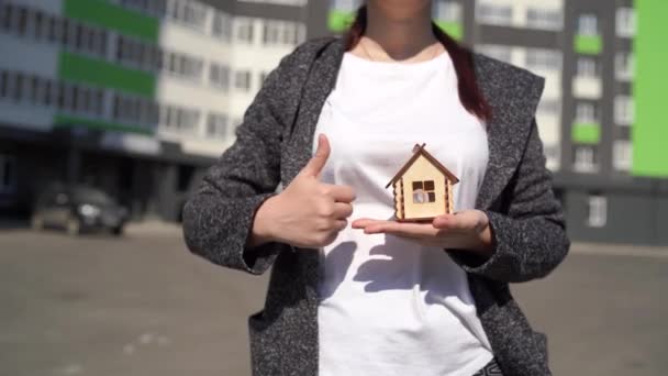 Close-up van de vrouw die kleine houten huis in de hand en andere hand tonen duim omhoog op de achtergrond van gebouwde huis. Concept van de aankoop van een nieuw appartement. — Stockvideo