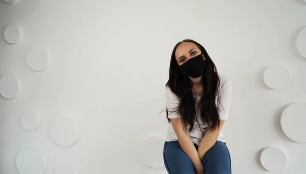 身穿黑色医疗面罩的年轻女子站在白墙的背景上 身穿白色T恤和蓝色牛仔裤的成年女性靠墙坐在椅子上 — 图库照片