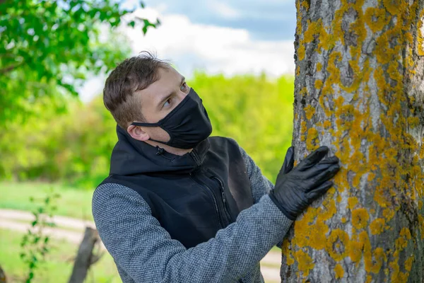 Молодий чоловік в чорній медичній масці і рукавичках обіймає дерево в лісі. Дорослий чоловік насолоджується природою в сільській місцевості в період епідемії коронавірусу . — стокове фото