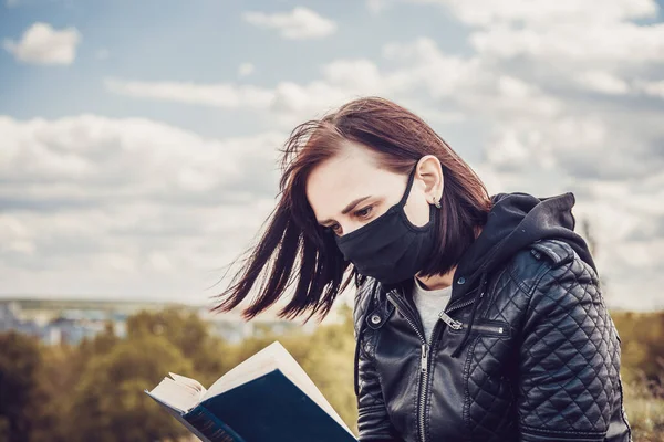 Großaufnahme einer jungen Frau in schwarzer medizinischer Maske und legerer Kleidung, die an bewölkten Tagen auf einem Hügel sitzt und Bücher liest. Erwachsene Frauen ruhen sich allein auf dem Land aus. — Stockfoto