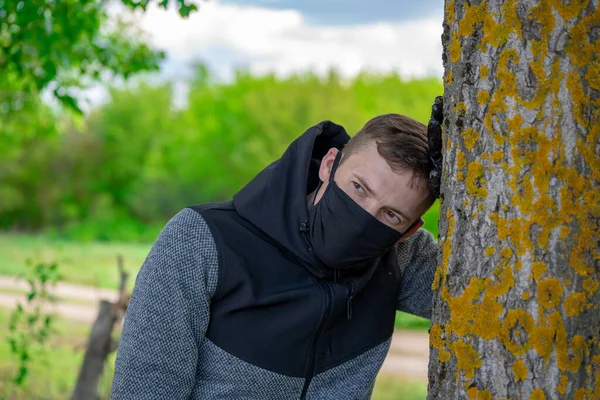 Junger Mann in schwarzer Arztmaske und Handschuhen lehnte im Wald an einem Baum. Erwachsene Männer genießen die Natur auf dem Land bei Coronavirus-Epidemie. — Stockfoto