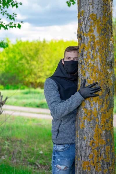 Ung man i svart medicinsk mask och handskar kramar träd i skogen. Vuxna män som njuter av naturen på landsbygden under perioden med epidemisk infektion orsakad av coronavirus. — Stockfoto