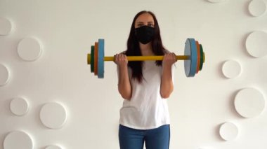 Siyah tıbbi maskeli genç bir kadın beyaz desenli duvara karşı renkli halteri kaldırıyor. Yetişkin kadın spor yapıyor, Coronavirüs salgını sırasında evde izole ediliyor..