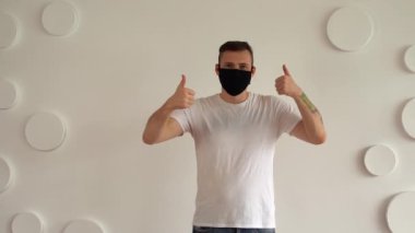 Siyah maskeli yakışıklı genç adam beyaz duvarın arka planında baş parmağını gösteriyor. Beyaz tişörtlü yetişkin erkek desenli duvara karşı onay işareti gösteriyor..