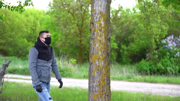 Hombre joven con máscara médica negra y guantes abrazando el árbol en el bosque. Hombre adulto disfrutando de la naturaleza en el campo en el período coronavirus infección epidémica . — Vídeo de stock