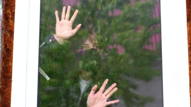 Mujer joven con máscara médica negra mirando por la ventana, poniendo las manos en la ventana aislada en casa durante la infección epidémica por coronavirus. Mujer pensativa con ojos marrones observando a través de la ventana . — Vídeo de stock