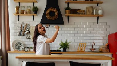 Aşırı neşeli, siyah saçlı, ağzı açık, elinde siyah kaşıkla modern mutfakta şarkı söylüyor gibi yapan bir kadın.