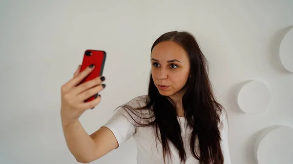 Jovem mulher tomando selfie no smartphone no fundo da parede branca estampada. Retrato de mulher passar o fim de semana em casa e fotografar de si mesma no celular . — Fotografia de Stock