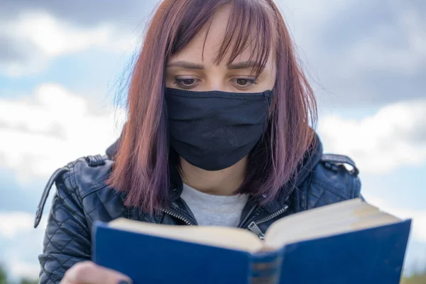 黒い医療用マスクとカジュアルな服を丘の上に座って 曇りの日に本を読んで若い女性の閉じる 田舎に一人でいる大人の女性 — ストック写真