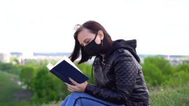 Siyah tıbbi maskeli ve günlük kıyafetleriyle tepede oturmuş bulutlu bir günde kitap okuyan genç bir kadın. Yetişkin dişi kırsalda tek başına dinleniyor..