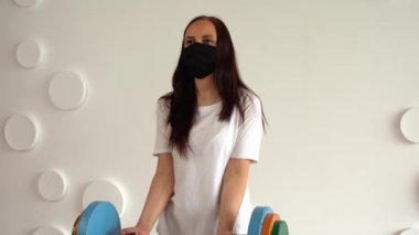Siyah tıbbi maskeli genç bir kadın beyaz desenli duvara karşı renkli halteri kaldırıyor. Yetişkin kadın spor yapıyor, Coronavirüs salgını sırasında evde izole ediliyor..