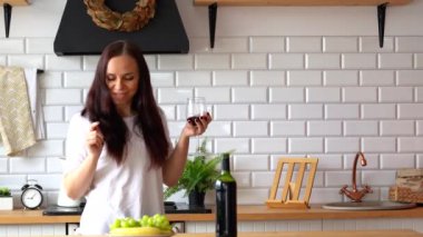 Genç bir kadın kırmızı şarap içip masada dans ediyor. Yetişkin bir kadının mutfakta bir bardak şarapla eğlenmesine yakın çekim.