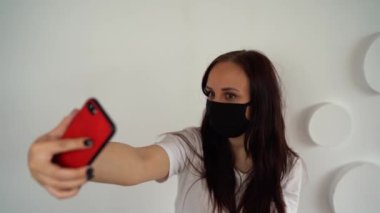 Siyah tıbbi maskeli genç bir kadın beyaz duvarın arka planında akıllı telefondan selfie çekiyor. Hafta sonunu evde geçiren bir kadının portresi ve cep telefonuyla kendi fotoğrafını çekmesi..