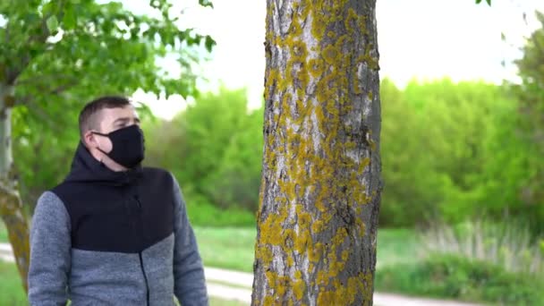 Jeune homme en masque médical noir et gants serrant l'arbre dans les bois. Homme adulte profitant de la nature à la campagne dans l'infection épidémique de coronavirus période . — Video