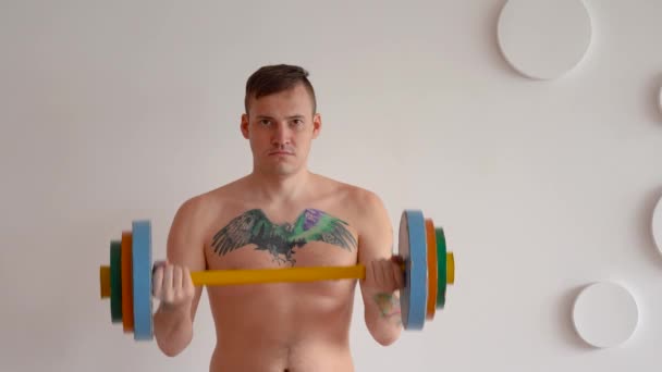 Young man with naked body and tattoo on chest lifting multicolored barbell against white patterned wall. Adult strong guy doing sport at home. — Stock Video