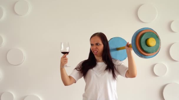 En ung kvinne med et glass vin i hånden og en mangefarget bjelle mot en hvit mønstervegg. Voksen dame som viser med sine følelser at dumbbell er bra, men vin er dårlig . – stockvideo