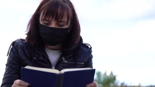 Gros plan de la jeune femme en masque médical noir et vêtements décontractés assis sur la colline et le livre de lecture le jour nuageux. Femme adulte se reposant seule dans la campagne. — Video