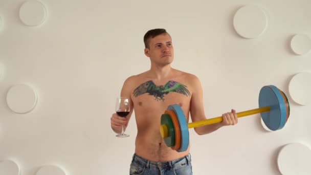 Young man with naked body and tattoo on chest lifting multicolored barbell and drinking red wine against white patterned wall. Adult strong guy doing sport and drinking alcohol. — Stock Video