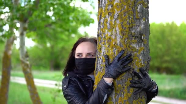 年轻的女人戴着黑色的医疗面罩 戴着手套 在林地里抱着树 成年女性在农村享受自然时期的大肠病毒感染 — 图库视频影像