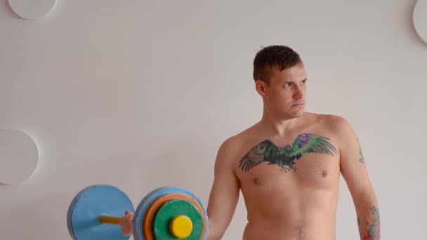 Giovane uomo con corpo nudo e tatuaggio sul petto sollevamento multicolore bilanciere contro parete fantasia bianca. Adulto ragazzo forte fare sport a casa . — Video Stock