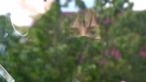 年轻女人戴着黑色的医疗面罩 看着窗外 在科罗纳韦病毒感染期间被隔离在家里 多愁善感的女性形象 棕色眼睛 透过窗户观察 — 图库视频影像