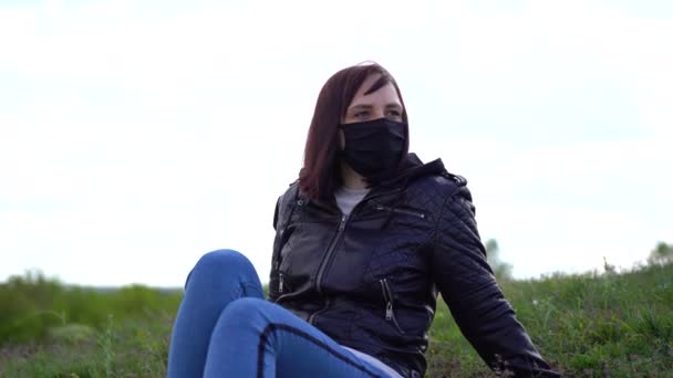 Jeune femme en masque médical noir et vêtements décontractés assis sur la colline par temps nuageux. Femme adulte se reposant seule, profitant de la nature à la campagne . — Video