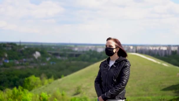 曇りの日に丘の上に黒い医療用マスクと革のジャケットの若い女性。田舎を一人で歩く大人の女性. — ストック動画